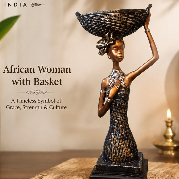 A decorative sculpture of an African woman carrying a basket on her head, displayed on a tabletop with a warm neutral background, plants, a woven mat, and a small brass lamp. Text on the left reads 'African Woman with Basket' and 'A Timeless Symbol of Grace, Strength & Culture.
