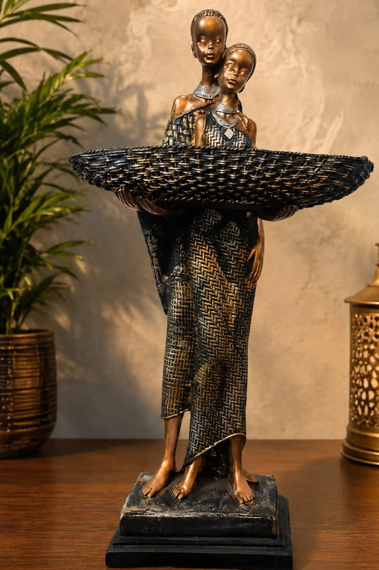 A bronze-finished decorative sculpture of two figures holding a large woven basket, displayed on a wooden surface against a textured beige wall, with a potted plant on the left and a lit brass lantern on the right.