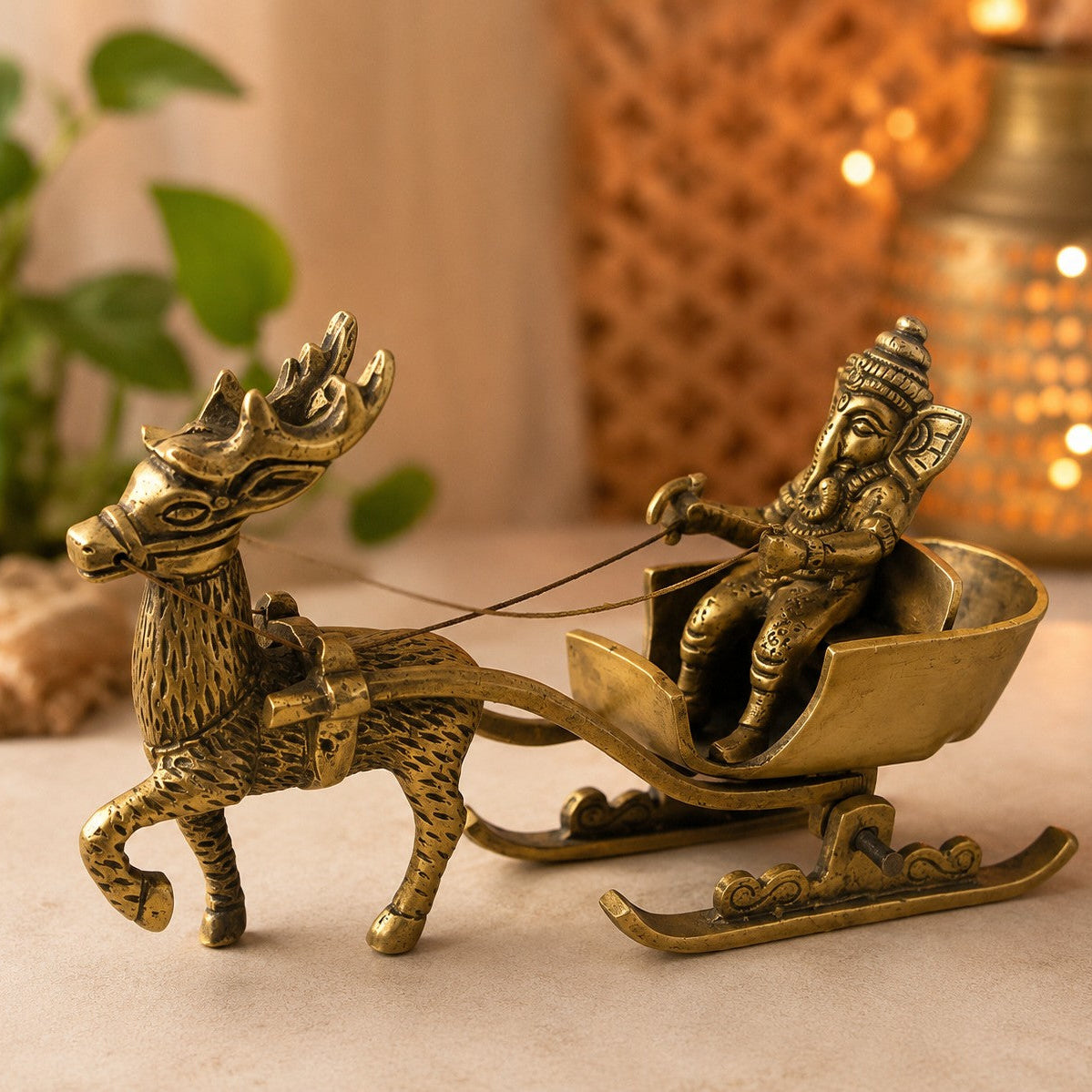 A brass decorative figurine of Ganesha seated in a sleigh pulled by a deer, displayed on a beige surface with warm bokeh background lights and green leaves.