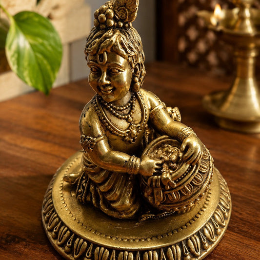 A small brass statue of a Krishna with his makhan matka , makhan chor  on a decorative round base, placed on a wooden surface with a blurred indoor background and green leaves in the upper left.